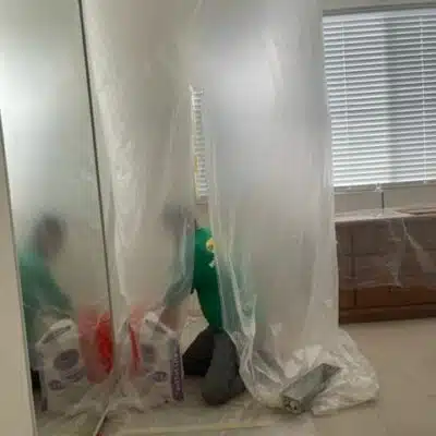 Worker in a green shirt behind a transparent plastic sheet, performing renovation or maintenance tasks in a residential bathroom, with tools and supplies visible.